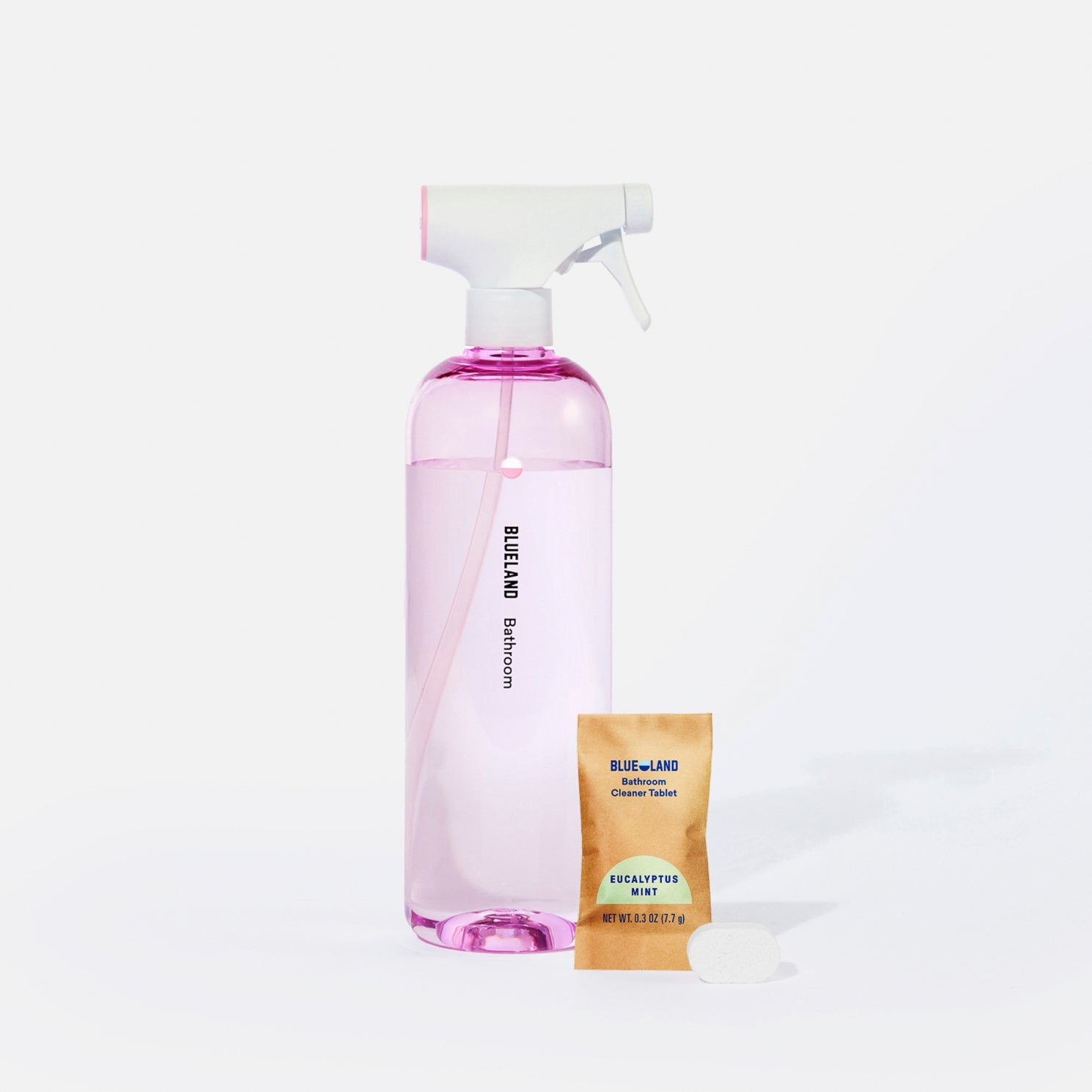 A clear spray bottle labeled Blueland Bathroom with pink liquid inside is next to a packet labeled Blueland Cleaner Tablet Eucalyptus Mint and a pink cleaning tablet on a white background.