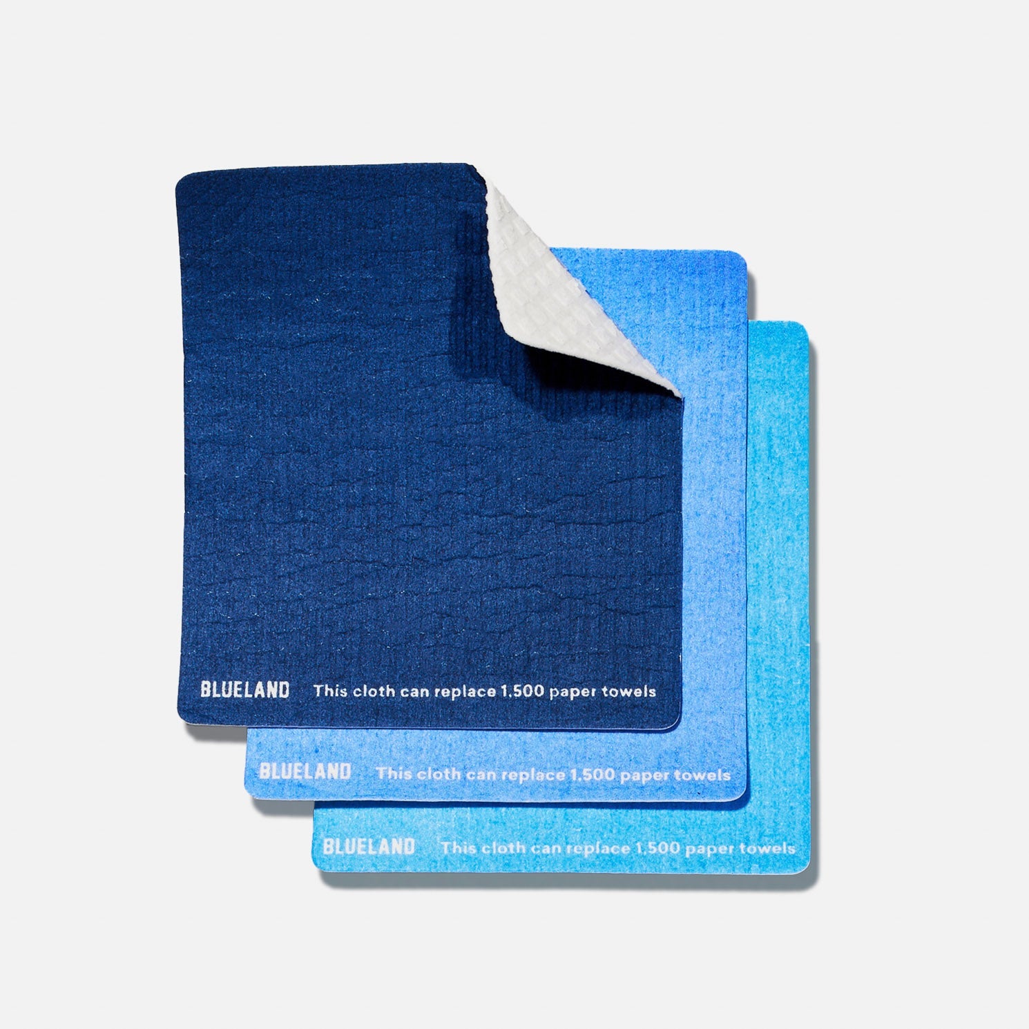 Three reusable cleaning cloths in varying shades of blue are stacked, with the top cloth’s corner folded to show its white underside. The cloths are branded Blueland and state they can replace 1,500 paper towels.