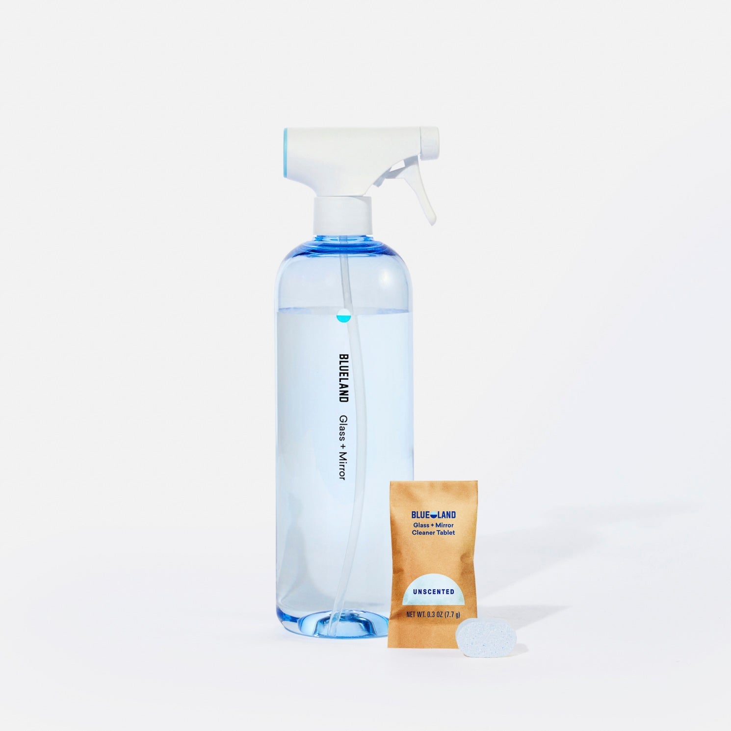 A clear spray bottle filled with blue liquid is labeled Blueland Clean and sits next to a brown packet labeled Blueland Cleaner Tablet Unscented and a white cleaning tablet, all on a white background.