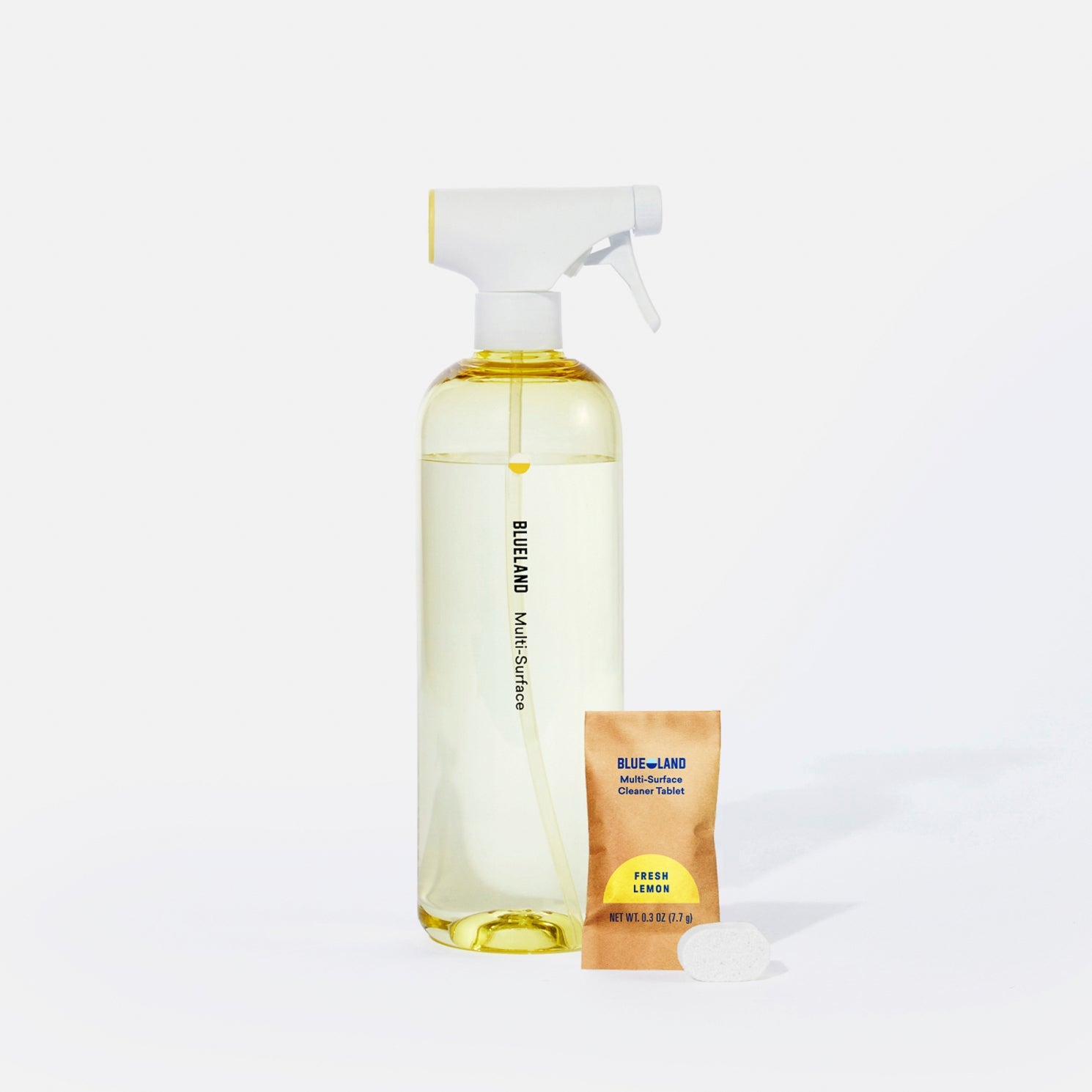 A clear spray bottle labeled Blueland Multi-Surface filled with yellow liquid sits next to a small packet labeled Blueland Fresh Lemon and a beige cleaning tablet, all on a white background.