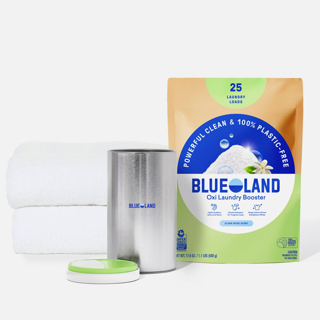 A package of Blueland Oxi Laundry Booster sits next to a pink canister with an open lid and two neatly folded white towels, all on a white background.