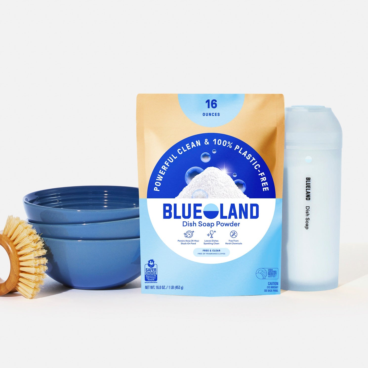 Blue bowls, a wooden dish brush, and a reusable bottle labeled Dish Soap sit beside Blueland’s Dish Soap Powder Starter Set—an eco-friendly, plastic-free cleaning powder in blue and tan packaging with a mountain graphic.