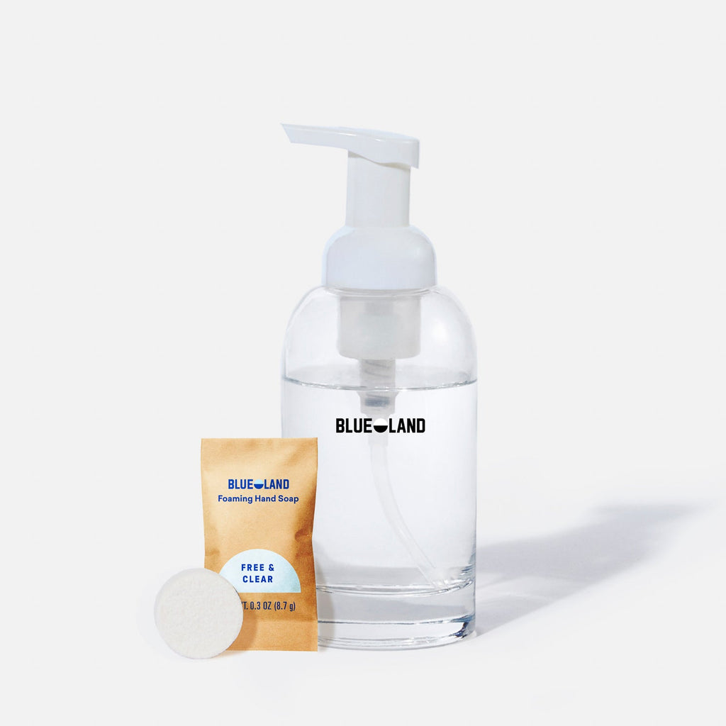 A clear pump bottle labeled Blueland is filled with liquid, next to a small packet labeled Blueland Foaming Hand Soap Free & Clear and a round white soap tablet, all on a white background.